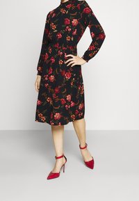 Robe noire à manches longues avec motifs floraux rouges et verts, taille cintrée, longueur au genou, assortie à des escarpins rouges brillants.