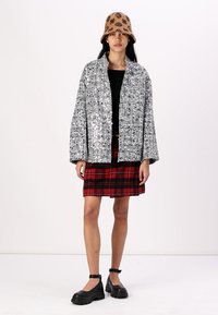 Veste oversize en sequins argentés, top court noir, jupe à carreaux rouges et chapeau seau à motifs marron, assortis avec des chaussures noires à semelles épaisses.