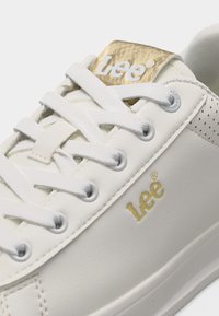 Λευκό αθλητικό παπούτσι Lee με λευκά κορδόνια, χρυσό λογότυπο στο πλάι και στη γλώσσα, και διάτρητη λεπτομέρεια κοντά στη φτέρνα.