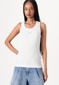 Hvit ribbestrikket tanktop med en liten beigefarget logo lapp i midten. Kombinert med lyseblå denimshorts med et gjentagende mønster.