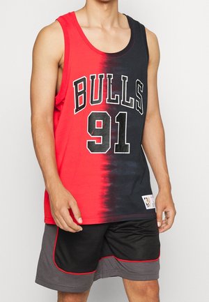 Mężczyzna w czerwono-czarnej bezrękawnikowej koszulce do koszykówki z nadrukiem "BULLS 91" z przodu oraz czarno-szortach do koszykówki z czerwonym obszyciem.
