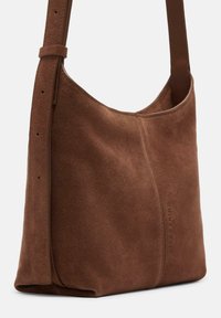 Brązowa zamszowa torba na ramię o gładkiej teksturze, zaokrąglonym kształcie i prostym designie. Posiada jeden pasek i subtelną wytłoczkę logo.