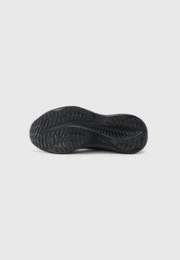Suela de zapatilla negra con goma texturizada y patrones de tracción variados. Diseño flexible con bordes suaves y una cresta central.