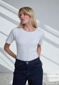 Femme blonde les mains derrière le dos, portant un haut blanc côtelé à manches courtes et un jean bleu foncé, debout sous une lumière douce.