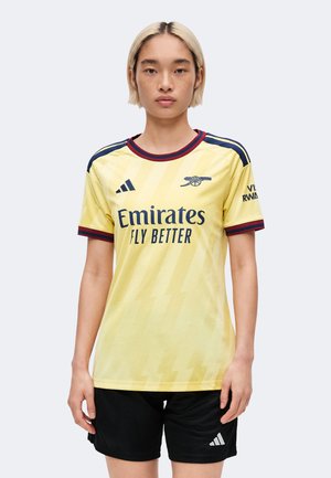 Ung person med kort blondt hår iført en gul Arsenal fodboldtrøje og sorte Adidas shorts, står oprejst mod en ensfarvet baggrund.