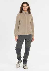 Fleece ritjack in lichtbruin met een gestructureerd patroon. Gecombineerd met donkergroene broek met zwarte accenten en zijzakken.