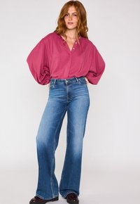 Blusa oversize bordeaux con scollatura a bottoni, abbinata a jeans a zampa d'elefante blu chiaro. Tessuto morbido e vestibilità rilassata con denim leggermente sbiadito.