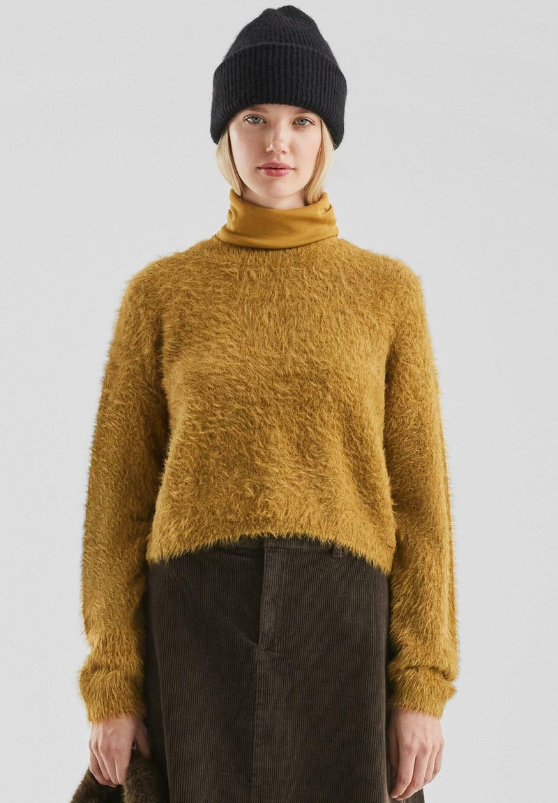Mosterdgele fuzzy trui met een ronde halslijn, lange mouwen en geribbelde manchetten; gecombineerd met een donkere corduroy rok en een zwarte gebreide beanie.