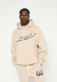 Felpa beige con tasca frontale e stampa grafica "MLTPLY Apparel" in nero. Abbinata a pantaloni della tuta beige coordinati con logo nero.