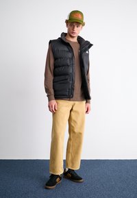 Gilet rembourré noir sur une chemise à manches longues marron, pantalon jaune à jambes larges et baskets noires avec des accents bruns ; casquette verte avec logo.