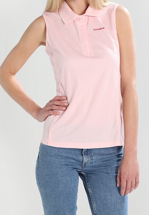 Femme portant un polo rose clair sans manches avec un col et un jean bleu, posant avec une main sur la hanche.