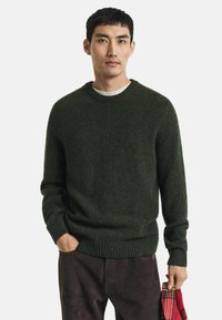Junger Mann in dunkelgrünem Strickpullover und dunklen Hosen, hält einen rot-schwarz karrierten Stoff in der rechten Hand, die linke Hand in der Tasche.