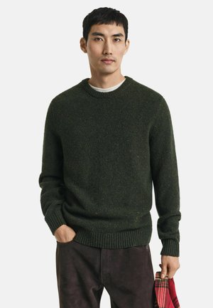 Young man wearing dark green knit sweater and dark pants, holding red and black plaid fabric in right hand, left hand in pocket.
