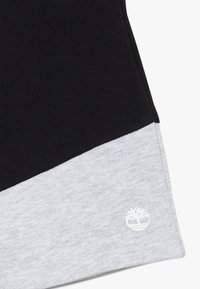 Musta puuvillakangas, jossa harmaa puuvillainen alaosa. Alareunassa vasemmalla pieni valkoinen puun logo. Sileä pinta ja yksinkertainen muotoilu.