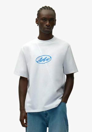 Giovane con capelli intrecciati che indossa una maglietta bianca a maniche corte con logo "Arte" blu e jeans blu, in piedi con una mano in tasca.