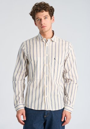 Mężczyzna z kręconymi włosami, ubrany w beżową, długim rękawem koszulę zapinaną na guziki z niebieskimi pionowymi paskami oraz niebieskie dżinsy, na jednolitym tle.