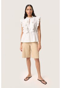 Hvid ærmeløs top med volanger, indsnævret talje og sløjfedetalje, parret med beige knælange shorts og sorte flip-flops.