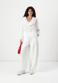 Blusa bianca con ruches e bottoni, abbinata a pantaloni bianchi larghi e sneakers bianche. Una borsa rossa è tenuta in una mano.