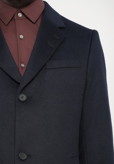 Marineblauer Woll-Blazer mit strukturiertem Revers, zwei Knöpfen und einer einzelnen Brusttasche, kombiniert mit einem burgunderfarbenen Hemd mit Knöpfen.