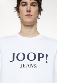 Felpa bianca con la scritta "JOOP! JEANS" stampata in bold con lettere blu navy. Collo rotondo classico e texture morbida e liscia.