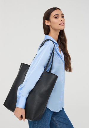 Borsa tote in pelle nera con texture liscia e doppi manici, tenuta accanto a una persona che indossa una camicia button-up azzurro chiaro e jeans.
