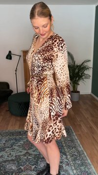 Ženska v živalskem vzorčnem obleki do kolen z dolgimi rokavi in črnimi gležnjaki, stoji na vzorčasti preprogi v dnevni sobi.