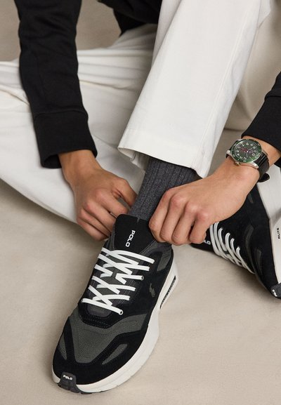 Personne portant un pantalon blanc et des chaussettes grises qui lacent des baskets Polo noires avec des lacets blancs en étant assise sur un sol clair.
