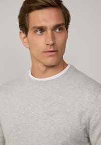 Suéter de punto gris claro con un escote redondo, textura suave y mangas largas, superpuesto a una camiseta blanca de cuello redondo.