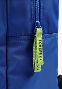 Mochila de tela azul con un acabado texturizado, con un tirador de cremallera verde etiquetado como "SCALPERS" y detalles de costura en contraste.