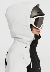 Chaqueta de nieve en blanco y negro con capucha, que cuenta con cierre de cremallera, cordón ajustable y bolsillo lateral, diseñada para deportes de invierno.