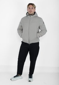 Giubbotto imbottito grigio con cappuccio, zip robusta e polsini elasticizzati. Indossato sopra pantaloni neri e sneakers grigie chiare, su uno sfondo bianco.