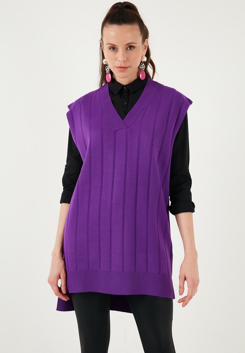 Purple knitted vest with a V-neck, ribbed texture, and sleeveless design. Worn over a black button-up shirt, with a long hem and side slits.