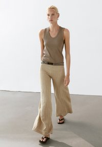 Beige mouwloze top gecombineerd met wijde gestreepte beige broek en zwarte sandalen. De outfit heeft een zwart met zilveren studs riem.