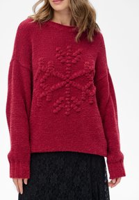 Czerwony, oversize'owy sweter z teksturowanym wzorem płatka śniegu na środku, z długimi rękawami i ściągaczami na mankietach, noszony na czarnej koronkowej spódnicy.