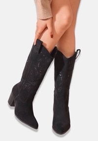 Bottes de cowboy en daim noir avec un bout pointu et un talon incliné. Présente des motifs floraux découpés complexes le long du tige et du bout.