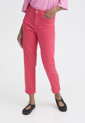 Nő rövid rózsaszín nadrágban, hozzáillő rózsaszín felsőben és fekete balettcipőben, egy kézzel a zsebében állva.