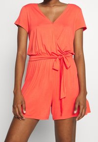 Mono corto de manga corta en color coral con escote en V, diseño envolvente y cinturón atado en la cintura, hecho de una tela suave con una textura lisa.