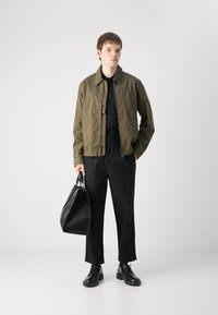 Olivengrønn lett jakke, svart polo-skjorte, tapered svarte bukser, svarte sko og en teksturert svart veske med håndtak. Enkle designelementer.