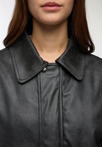 Giacca di pelle nera con un ampio colletto, dotata di chiusura con bottoni e texture liscia, che mette in evidenza cuciture distintive lungo la parte anteriore.