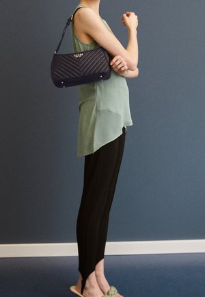 Personne portant un haut vert sans manches, des leggings noirs et des sandales vertes, avec un sac à bandoulière matelassé noir de la marque Guess sur le bras.