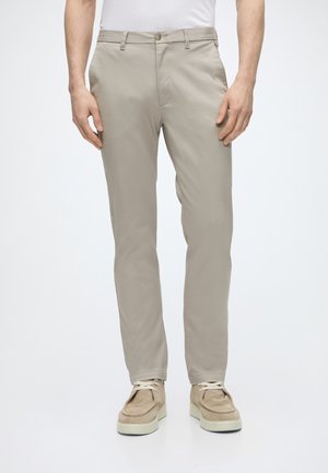 Hombre con pantalones ajustados beige, camisa blanca y zapatos casuales beige con cordones, de pie contra un fondo liso.