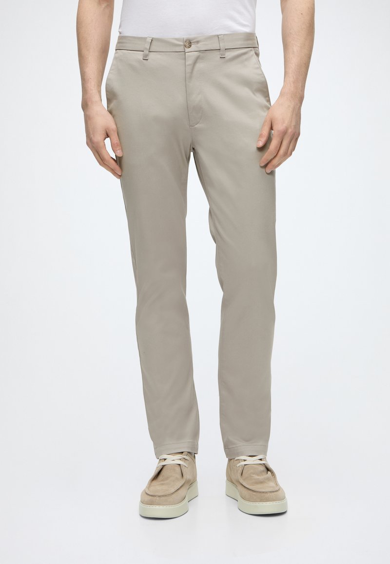 Άνδρας που φοράει μπεζ παντελόνια slim-fit, λευκό πουκάμισο και μπεζ casual δερμάτινα παπούτσια με κορδόνια, στέκεται μπροστά σε απλό φόντο.