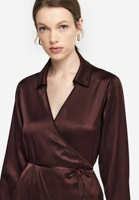 Burgundy satin inslaget blus med långa ärmar, en skuren krage och en knytning vid midjan. Slät textur med lätt glans.