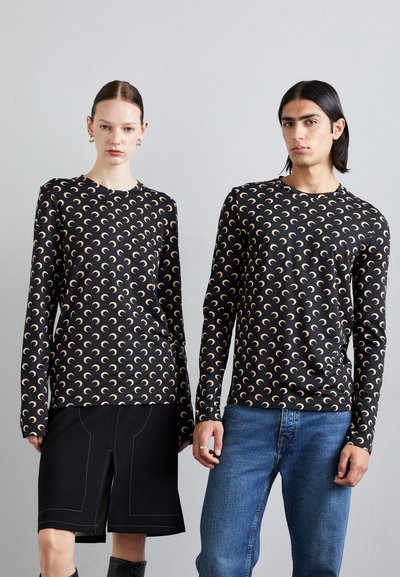 Langärmlige schwarze Hemden mit einem cremefarbenen Halbmondmuster, getragen von zwei Models, kombiniert mit einem schwarzen Rock und blauen Jeans.