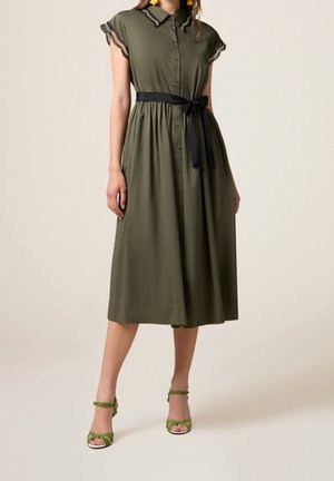 Napitettava mekko oliivinvihreänä, lyhyillä kapeilla hihoilla ja pitsireunuksella, vyötärö nauhavyöllä korostettuna, yhdistetty vihreisiin remmikenkiin.