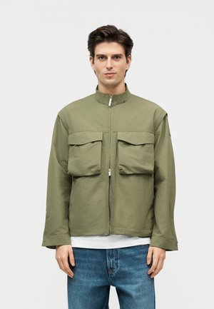 Mees, kellel on seljas oliiviroheline lukuga jakk kahe suure esitaskuga ja sinised teksad, seisab tavalise heleda tausta ees.