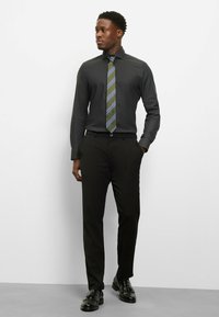 Schwarzes Hemd mit langen Ärmeln, kombiniert mit schwarzer Hose und glänzenden schwarzen Lederschuhen. Die Krawatte hat grüne und violette Streifen.