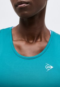 Prim-plan cu gâtul și partea superioară a pieptului unei persoane care poartă un top sportiv verde-albăstrui, cu un mic logo alb pe partea dreaptă.