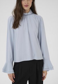 Žena v světle modré halence s vysokým límcem, na ramenech s nařasením a s rozšířenými manžetami, v kombinaci s černými skládacími kalhotami.