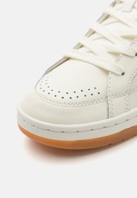 White leather sneaker with perforated toebox, flat laces, and a cushioned, light brown rubber sole. Stitching accents along the design.